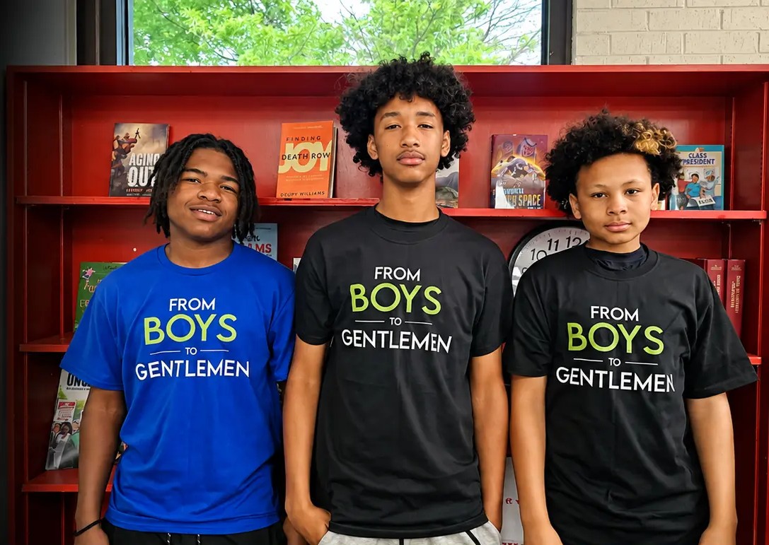 Three students in From Boys to Gentlemen shirts in front of a school bookshelf.